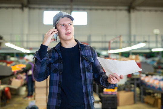 Focused strict young supervisor standing with papers in hands in citrus sorting workshop of agricultural factory, solving work issues on phone ..