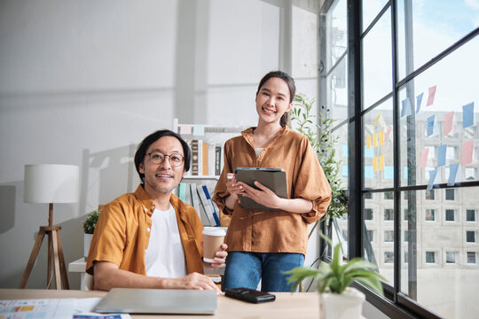 Portrait of two Asian startup entrepreneur partners smiling and looking at camera in casual small business office, creative marketing jobs, E-commerce freelance work, happy and cheerful coworker team.