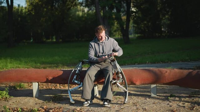 Young man in gray hoodie sitting on park bench adjusting metallic spring stilts on sunny day, surrounded by green grass and trees, with focused expression in peaceful outdoor setting