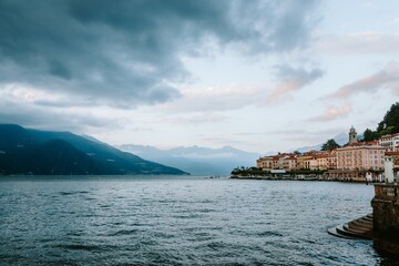 Lake como and the beautiful village of bellagio reflecting on the water under a cloudy sky with mountains in the background, creating a dramatic and picturesque landscape