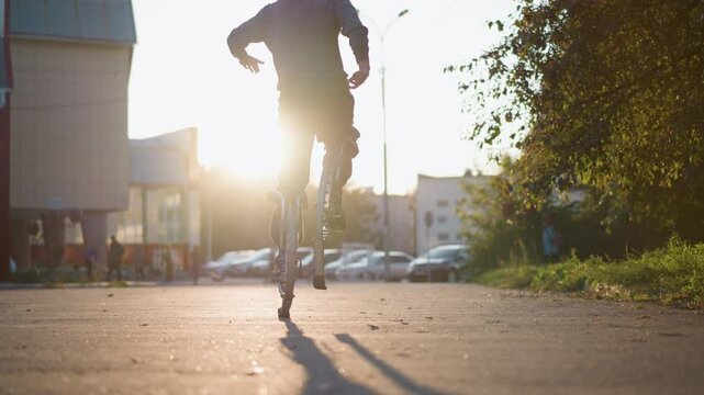Back shot of guy wearing gray hoodie and dark gray pants jumping on spring stilts on paved road in residential area surrounded by houses during sunset, showcasing dynamic motion