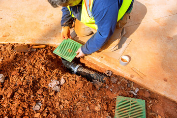 Construction worker wearing safety vest is installing drainage grates over pipes in trench ground around is covered with red soil, debris, indicating ongoing work.