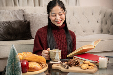Asian woman in red sweater sitting reading a book and sipping a warm drink in living room during...