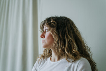 Profile of a thoughtful young woman with curly hair, gazing towards a window with a white curtain,...