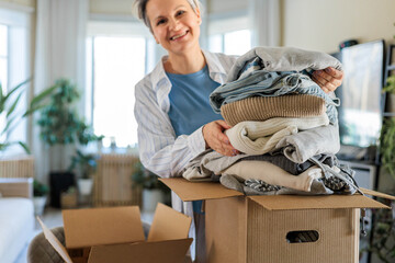 Portrait of smiling gray haired mature woman pack clothes pile to cardboard box for donation