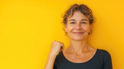 Smiling middle-aged woman raising a clenched fist with rolled-up sleeve against a bright yellow background, conveying empowerment and positivity.