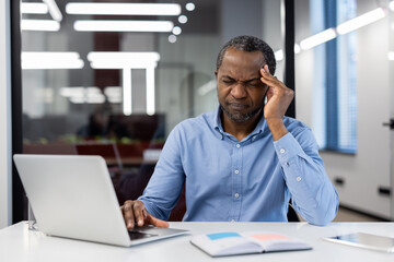 Mature African American businessman experiences stress at office desk, working on laptop. Hand on...