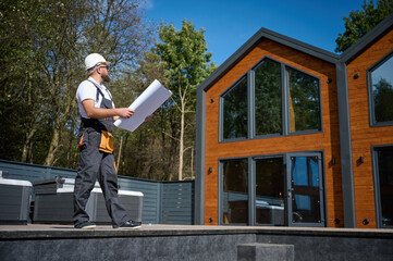 Architect hold plan. Bearded architect wearing hardhat holding project blueprints paper plans standing near comlex of new modern houses looking to something at construction site.
