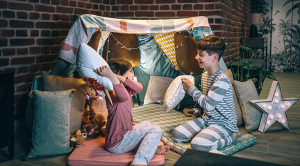 Funny children having fun playing pillow fight on handmade shelter tent in living room. Excited boy...