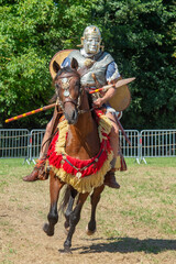 chevalier en armure avec sa lance et son casque sur un cheval brun rev&ecirc;tu d'un costume rouge et or 