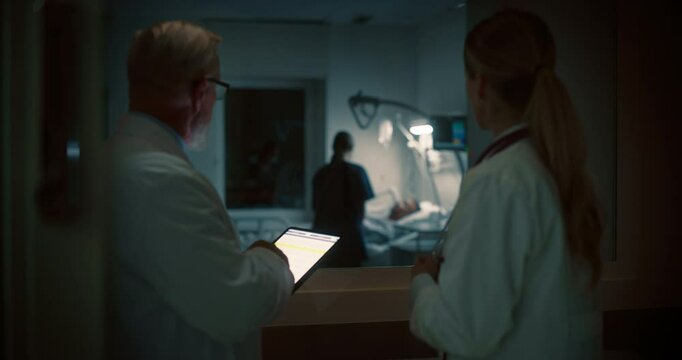 Senior Doctor Holding a Tablet Computer, Sharing Critical Patient Data With a Female Colleague Outside a Hospital Ward with Nurse and Patient, Their Discussion Focused on Treatment Strategies