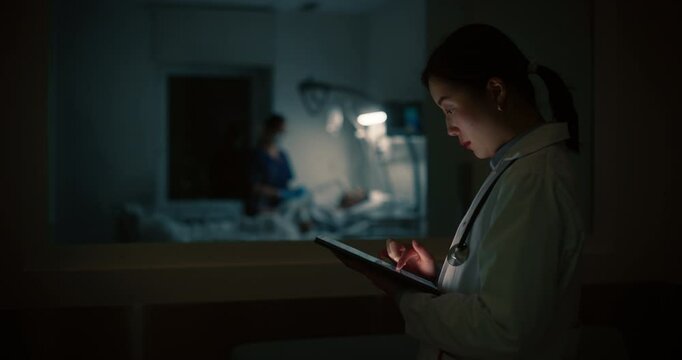 Asian Female Physician Standing in the Dark Hallway Outside a Hospital Intensive Care Ward, Focused on a Tablet Computer while a Nurse Taking Care of a Resting Patient in the Background