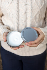 A person in a cozy sweater holds an open container of cream outside, showcasing a product ready for use in a natural, serene environment, emphasizing relaxation.
