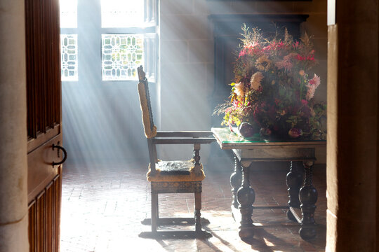 Sunlit Elegance &ndash; A charming scene featuring an old wooden chair beside a rustic table adorned with a delicate flower arrangement. The warm, bright light streaming through the window creates a serene 