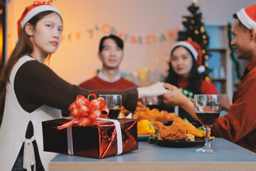 Group of young Asian man and women as friends having fun at a New Year's celebration, holding gift boxes standing by Christmas tree decoration, midnight countdown Party at home with holiday season.