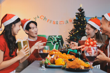 Group of young Asian man and women as friends having fun at a New Year's celebration, holding gift boxes standing by Christmas tree decoration, midnight countdown Party at home with holiday season.