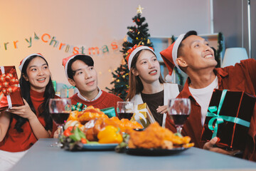 Group of young Asian man and women as friends having fun at a New Year's celebration, holding gift boxes standing by Christmas tree decoration, midnight countdown Party at home with holiday season.