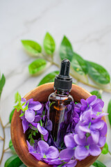 A mortar with purple flowers and an amber vial full of essential oil, on a marble surface. Close-up image