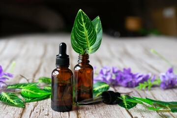 Amber vial of essential oil with a dropper, and another with leaves in it, on a wooden surface, with purple flowers and leaves in the background