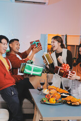Group of young Asian man and women as friends having fun at a New Year's celebration, holding gift boxes standing by Christmas tree decoration, midnight countdown Party at home with holiday season.