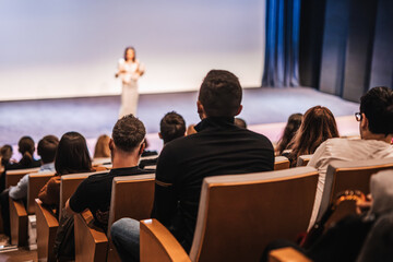 Business and entrepreneurship symposium. Female speaker giving a talk at business meeting. Audience in conference hall. Rear view of unrecognized participant in audience.