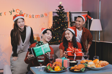 Group of young Asian man and women as friends having fun at a New Year's celebration, holding gift boxes standing by Christmas tree decoration, midnight countdown Party at home with holiday season.