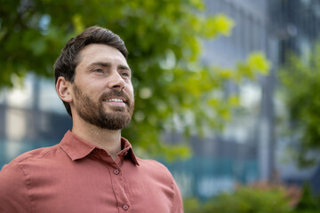 Mature businessman wearing casual shirt smiling outdoors with green leaves and office buildings...