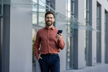 Mature businessman walking outside, holding smartphone, displaying confidence and professionalism. Wearing smart casual attire with modern architecture. Represents success, technology, communication.