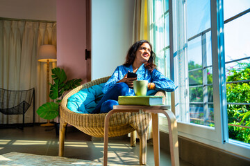 Happy Indian woman on video call with coffee mug, sitting by sunlit window, enjoying cozy morning