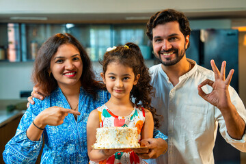 Happy Indian couple with their daughter holding a cake, sharing smiles and joy in the kitchen
