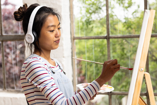 asian female teenager painting on canvas at home, wearing headphones, enjoying creative time