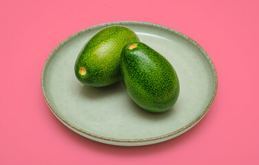 Two vibrant green avocados rest on a soft green ceramic plate. Placed against a bright pink background, creating a colorful and modern composition.