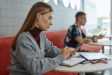 Side view of concentrated woman using phone for remote work or studying while sitting at table in...