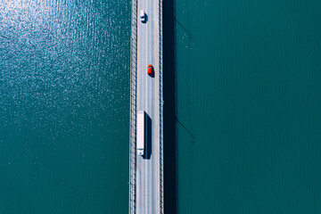 Aerial drone view of large freight transporter semi-truck and cars driving on the bridge road over blue sea lake river