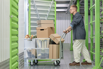 Full length shot of handsome man loading cart with cardboard boxes into self storage unit at...
