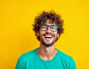 Candid portrait of a handsome curly-haired young man in a turquoise t-shirt and glasses on a yellow background laughing.