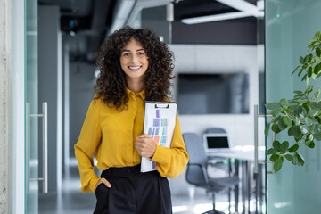 Confident businesswoma holding clipboard in modern office with plants and open laptop, presenting...
