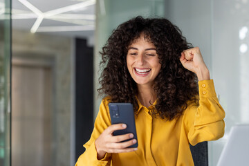 Excited woman celebrating success using smartphone in modern office. Happy expression, closed fist...