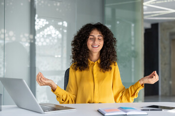 Woman meditates at office desk, demonstrating mindfulness with laptop and phone nearby. She is...