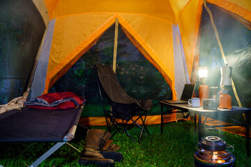 Camping tent, folding bed inside the tent at night in the forest, gas lantern and coffee set,...