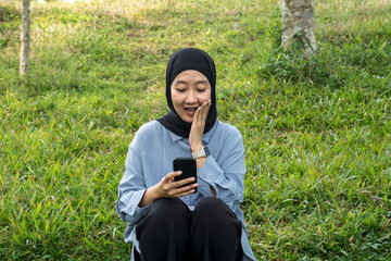 Happy Woman in Hijab Sitting on Grass Holding Smartphone