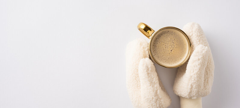 Hands wearing warm mittens hold a gold-rimmed coffee cup, creating a cozy winter atmosphere. The minimal white background emphasizes the warmth and comfort of the scene.