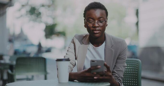 Smiling young woman joyful african american chatting in social media drinking coffee sitting at cafe outdoors. Application. Happy, emotional, urban, business, cellphone, face, relaxed. Slow motion