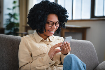 Smiling African woman using cell phone relaxing on couch at home. Happy lady sitting on sofa...