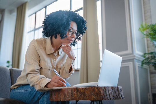 Young concentrate black woman using laptop at home and taking notes, happy african american female working remotely with computer while sitting on couch in living room at home