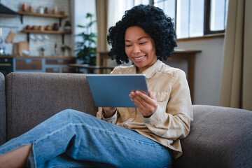 Close up of smiling young African woman relaxing on sofa using tablet computer reading book...