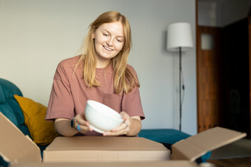 Young 30s woman opening parcel with plates indoors. Happy millennial female client satisfied with ordered purchase. Fast delivery service, online shopping concept