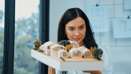 Close up of skilled young beautiful caucasian architect lift house model left side and right sight to check and focus on inspecting mistake point at modern office with blueprint. Immaculate.