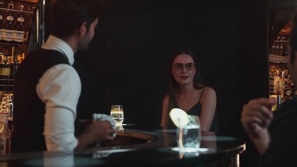 Focusing on beautiful woman guest sitting in special drink gin fizz at luxurious counter bar at vibrant nightlife party atmosphere. Smart bartender wiping glass while talking with customer. Vinosity.