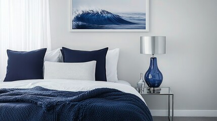 A minimalist blue and white bedroom with a sleek navy throw blanket over white bedding, complemented by art prints of ocean waves. A silver lamp and blue vase add modern elegance to the side table.